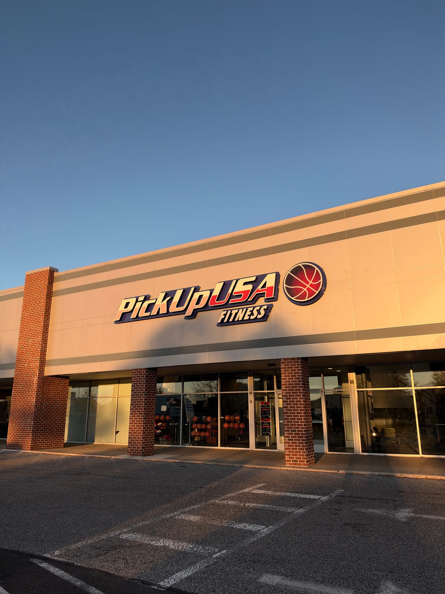 Pick up USA Fitness storefront with large sign and basketball logo, brick columns, glass doors, and a clear blue sky; warm sunlight casting long shadows on the facade.