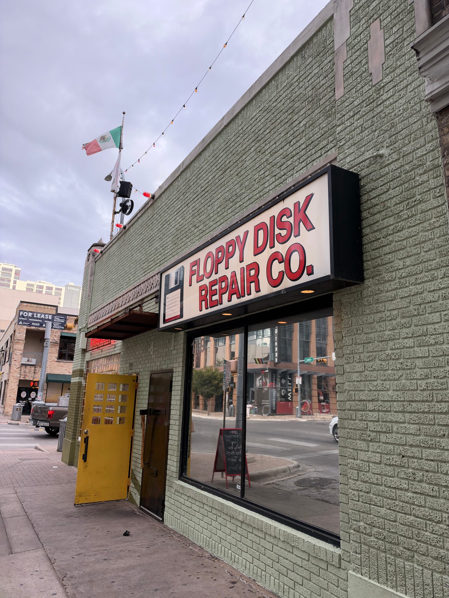 Floppy Disk Repair Co. storefront on a light green brick building with a large sign above the window; a yellow door is propped open at the side, and string lights stretch above the street. An American flag and a flag are on a pole atop the building.