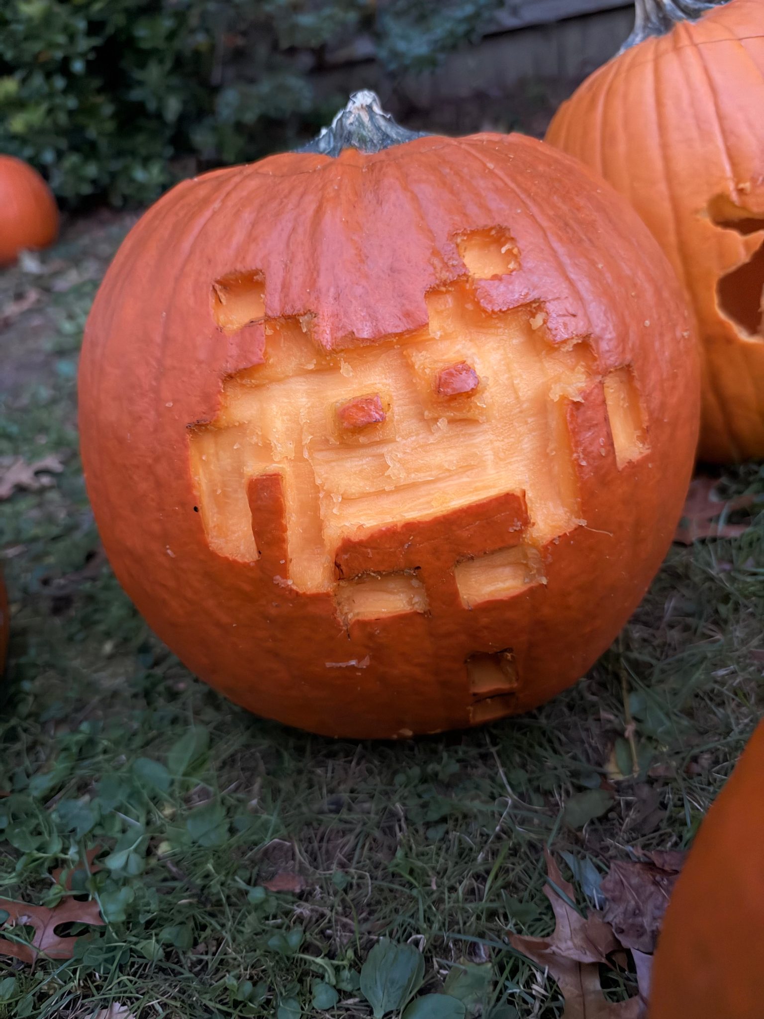 A carved pumpkin on the grass with a geometric, pixelated face cut into the front, unevenly carved with square and rectangular gaps; another pumpkin is partly visible to the right.