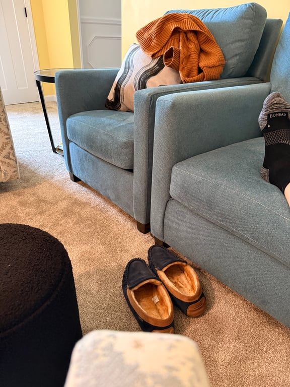 blue easy chair with a foot resting on it shod in a black sock. In front of the chair on the rug rests two navy blue slippers